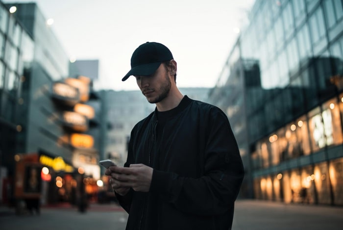 En mann med sort caps og genser som står i en gate og trykker på en telefon